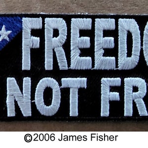 May include: A black patch with white embroidered text that reads "FREEDOM IS NOT FREE". A blue patch with white stars is in the upper left corner.