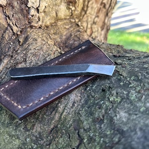 May include: A small, dark metal chisel rests on a brown leather sheath with white stitching. The chisel has a beveled edge and a flat top. The leather sheath is rectangular and appears to be handmade.