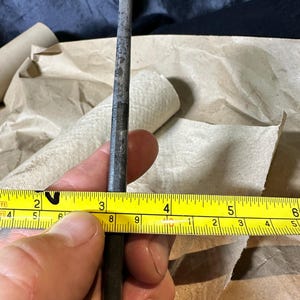 Puede incluir: Una herramienta de metal, de aproximadamente 7,6 cm de largo, se sostiene junto a una cinta m&eacute;trica amarilla. La herramienta tiene un aspecto oscuro y desgastado. La cinta m&eacute;trica muestra medidas en pulgadas.