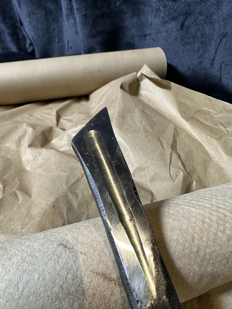Puede incluir: Primer plano de un cincel de carpinter&iacute;a met&aacute;lico con un borde biselado. El cincel tiene un acabado met&aacute;lico oscuro con una superficie pulida y reflectante. El fondo incluye papel marr&oacute;n y un tubo de cart&oacute;n enrollado.