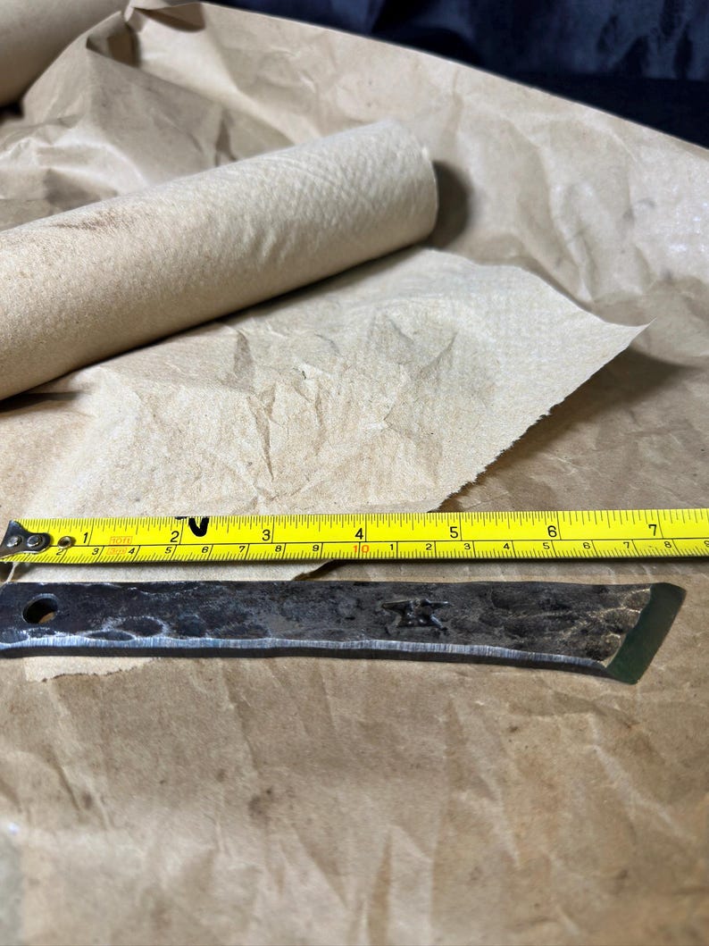 Puede incluir: Una hoja de metal forjada a mano con una cinta m&eacute;trica amarilla y un rollo de toallas de papel marr&oacute;n. La hoja mide aproximadamente 15 cm de largo. La cinta m&eacute;trica muestra pulgadas y fracciones de pulgada.