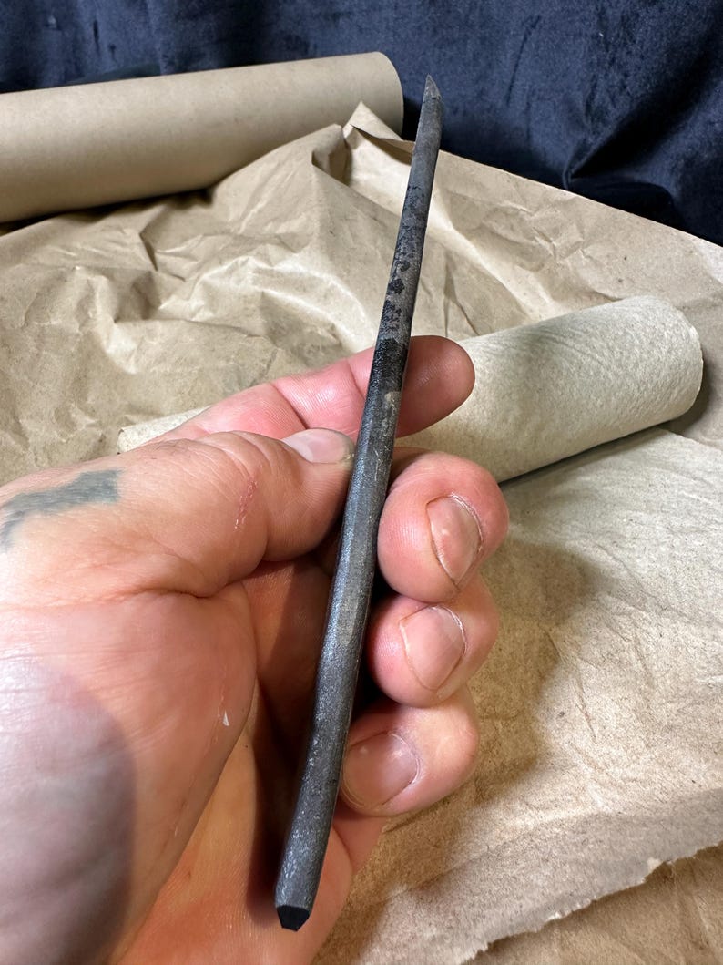 Puede incluir: Una herramienta met&aacute;lica puntiaguda de color gris oscuro con una punta negra. La herramienta se sostiene en una mano, con papel marr&oacute;n y tela oscura en el fondo.