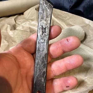 Puede incluir: Herramienta de metal forjada a mano con una superficie texturizada y un emblema en forma de estrella. La herramienta tiene un borde biselado y un agujero en la base. El metal tiene un aspecto oscuro y envejecido.