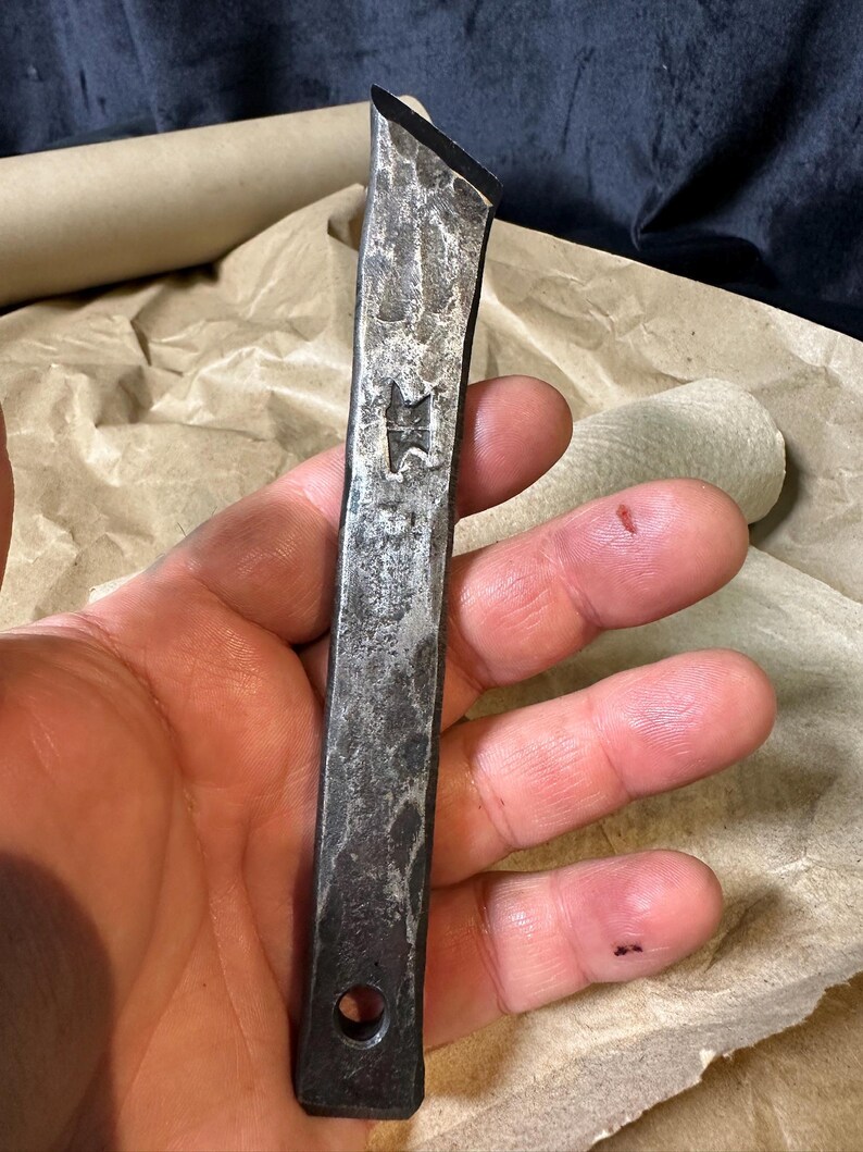 Puede incluir: Herramienta de metal forjada a mano con una superficie texturizada y un emblema en forma de estrella. La herramienta tiene un borde biselado y un agujero en la base. El metal tiene un aspecto oscuro y envejecido.
