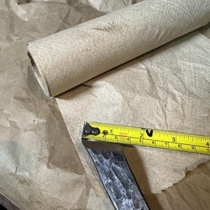 Puede incluir: Un cincel de metal con una cinta m&eacute;trica amarilla que muestra una medida de aproximadamente 7,6 cm. Un rollo de papel marr&oacute;n enrollado est&aacute; en el fondo, descansando sobre una hoja de papel marr&oacute;n.