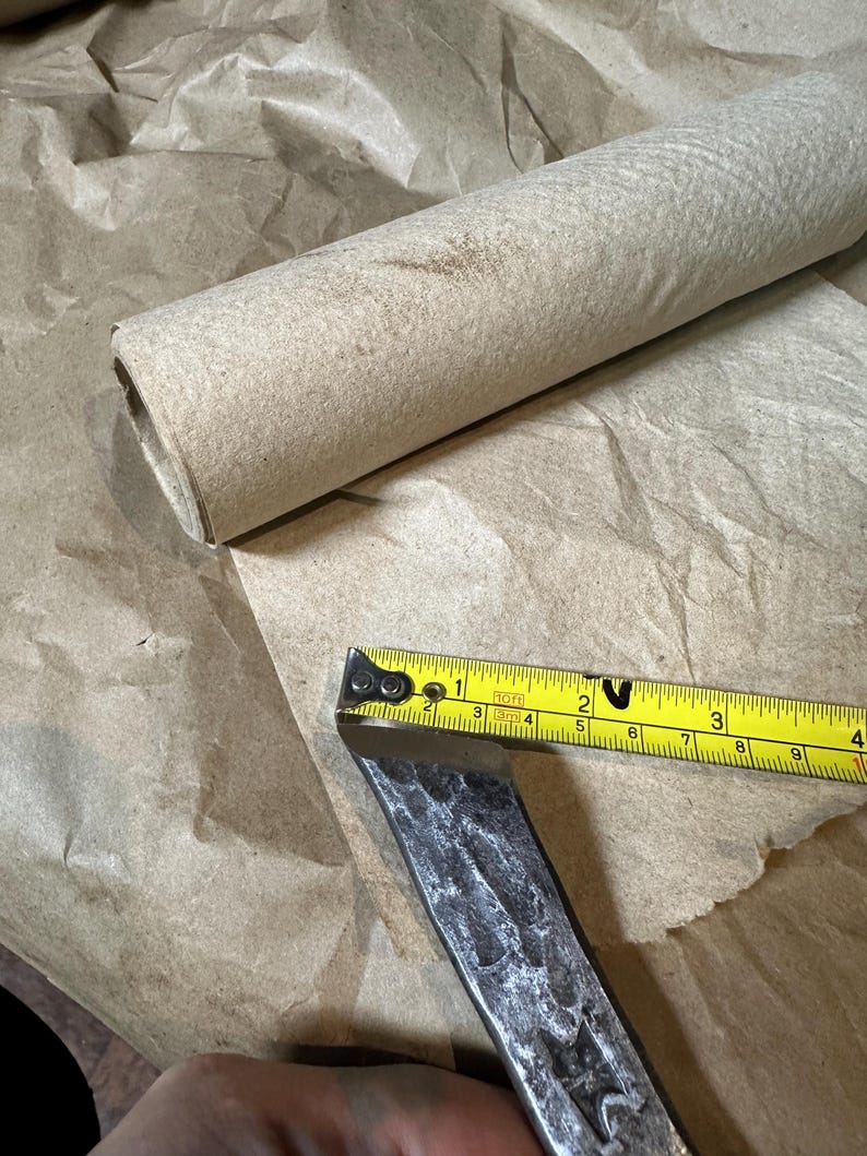 Puede incluir: Un cincel de metal con una cinta m&eacute;trica amarilla que muestra una medida de aproximadamente 7,6 cm. Un rollo de papel marr&oacute;n enrollado est&aacute; en el fondo, descansando sobre una hoja de papel marr&oacute;n.