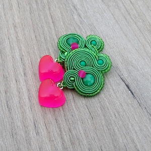 May include: A handmade green soutache brooch with a swirling design, accented with green and pink beads. Two bright pink heart-shaped charms dangle from the brooch. The brooch is set against a wooden background.