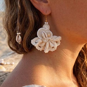 Peut inclure: Une boucle d'oreille pendante de couleur crème composée d'un coquillage cauri central entouré de cordons enroulés et d'une bordure en dentelle en forme d'éventail. Elle est suspendue à un crochet doré et ornée de petites perles blanches.
