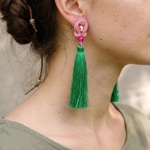 May include: A long dangle earring featuring a pink soutache braided top with a central clear crystal, a small pink bead, and a long, vibrant green silk thread tassel hanging from the bottom. The earring is secured with a post-style backing.