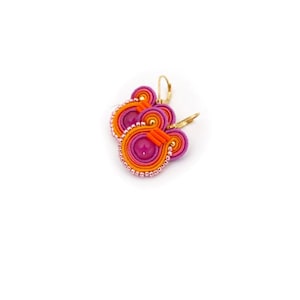Puede incluir: Un par de pendientes con un diseño de espiral rosa y naranja y una pequeña perla rosa en el centro. Los pendientes tienen ganchos dorados.