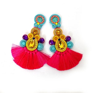 Pode incluir: Um par de brincos de borla rosa brilhante com detalhes de contas coloridas. Os brincos apresentam um grande cristal em forma de l&aacute;grima no centro, rodeado por contas menores em tons de azul, amarelo e roxo.