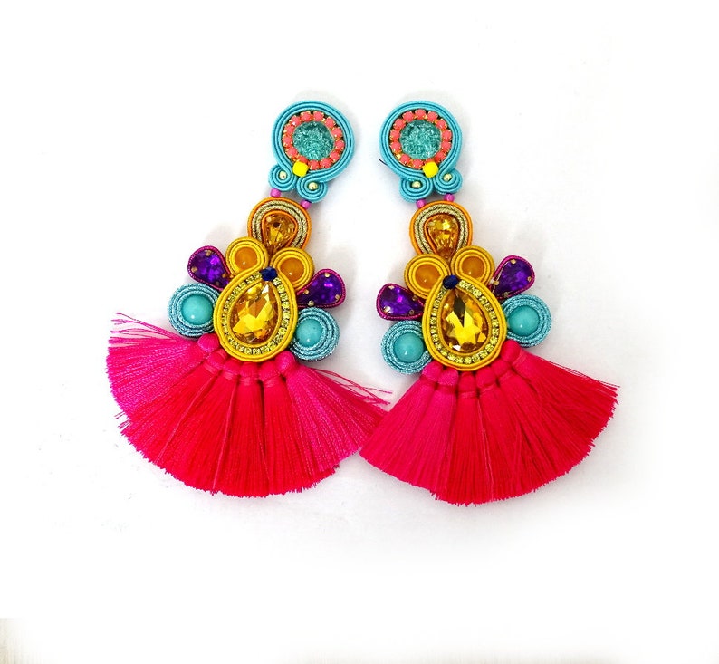 Pode incluir: Um par de brincos de borla rosa brilhante com detalhes de contas coloridas. Os brincos apresentam um grande cristal em forma de l&aacute;grima no centro, rodeado por contas menores em tons de azul, amarelo e roxo.