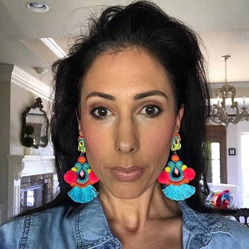 A pair of large, colorful dangle earrings with turquoise, pink, yellow, green, and orange tassels. The earrings have a beaded design with a pink pom-pom at the top.