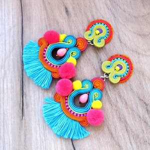 A pair of colorful earrings with blue fringe, pink pom poms, yellow and green swirls, and orange and pink beads.