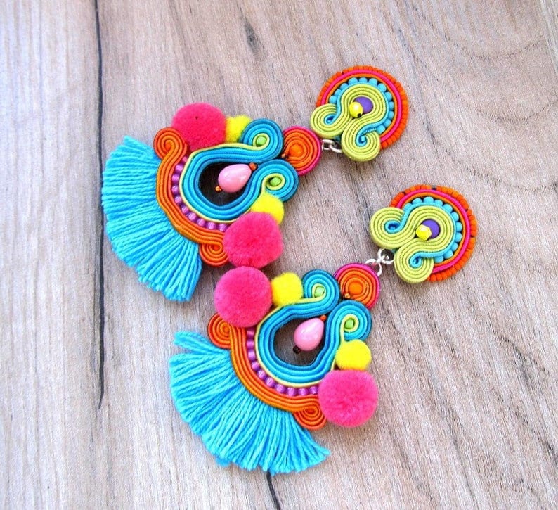 A pair of colorful earrings with blue fringe, pink pom poms, yellow and green swirls, and orange and pink beads.