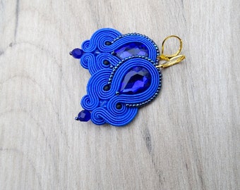 May include: A pair of blue earrings with a teardrop-shaped crystal in the center. The earrings are made of blue thread and have a gold hook.