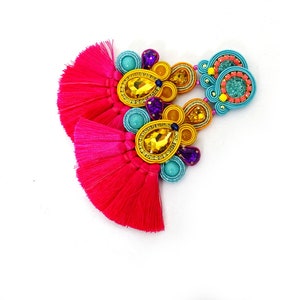 Pode incluir: Um par de brincos de borla rosa com enfeites de contas e fio coloridos. Os brincos t&ecirc;m detalhes em turquesa, amarelo e roxo.