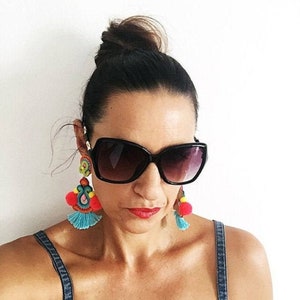 A pair of large, black, cat-eye sunglasses with dark lenses. The sunglasses are worn by a person with dark hair pulled back in a bun. The person is wearing large, colorful earrings with pink, blue, yellow, and green pom-poms.