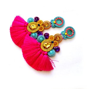 Pode incluir: Um par de brincos coloridos com borlas rosa, contas turquesa e amarelas e detalhes dourados.