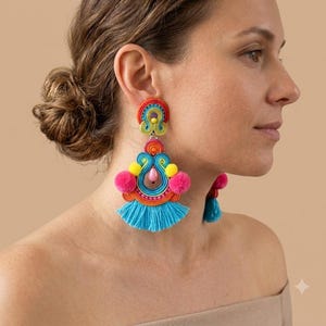 Pode incluir: Brincos pendentes com um design soutache multicolorido em fios vermelhos, laranja, amarelos e azul brilhante. Os brincos incluem pequenos pompons cor-de-rosa, uma conta central em forma de l&aacute;grima rosa e franjas azuis na base.