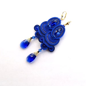 Puede incluir: Un par de pendientes de soutache azules hechos a mano con detalles dorados. Los pendientes presentan un colgante de cristal azul en forma de lágrima y un gran cristal azul ovalado. Cierre de palanca dorado.