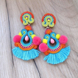 A pair of colorful dangle earrings with blue tassels, pink pom poms, and yellow pom poms. The earrings are made of woven threads in shades of blue, green, yellow, orange, and pink. The earrings have a silver hook closure.