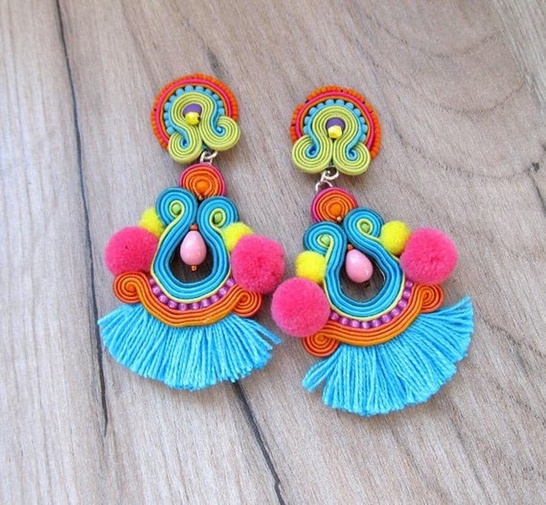 A pair of colorful dangle earrings with blue tassels, pink pom poms, and yellow pom poms. The earrings are made of woven threads in shades of blue, green, yellow, orange, and pink. The earrings have a silver hook closure.