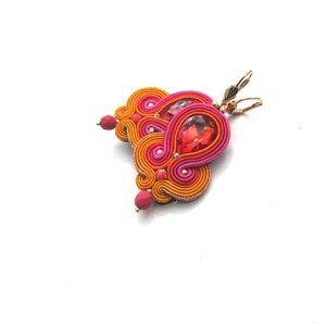 Könnte beinhalten: Ein Paar Ohrringe mit einem tropfenförmigen roten Edelstein, umgeben von orangefarbenem und rosafarbenem Garn. Die Ohrringe sind aus goldfarbenem Metall gefertigt.