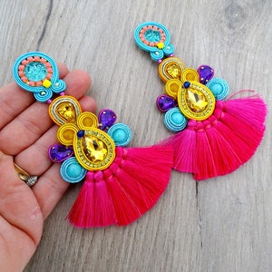 Pode incluir: Um par de brincos coloridos com borlas rosa. Os brincos s&atilde;o feitos com uma variedade de contas e pedras brilhantes em tons de azul, amarelo e roxo.