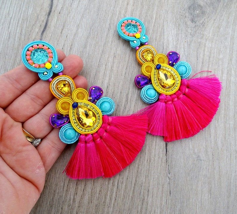 Pode incluir: Um par de brincos coloridos com borlas rosa. Os brincos s&atilde;o feitos com uma variedade de contas e pedras brilhantes em tons de azul, amarelo e roxo.