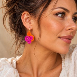 May include: A dangling earring featuring a vibrant design of coiled orange and bright pink cord, accented with small matching beads. The earring is suspended from a simple gold-toned metal hook, creating a layered, swirling pattern.