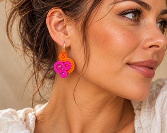 Puede incluir: Un pendiente colgante con un diseño vibrante de cordón enrollado en color naranja y rosa brillante, acentuado con pequeñas cuentas a juego. El pendiente cuelga de un sencillo gancho de metal dorado, creando un patrón de espirales superpuestas.
