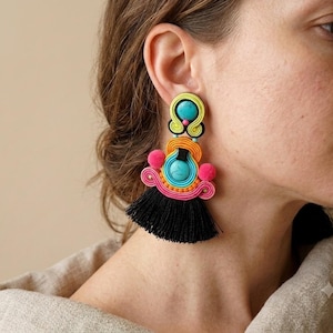 May include: A statement earring featuring a turquoise stone set in lime green cord, accented with orange and pink cord loops, small pink pom-poms, and a large black tassel at the base. The design incorporates intricate soutache embroidery techniques.