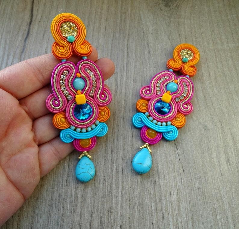 May include: A pair of long, colorful earrings with turquoise stones. The earrings are made of pink, orange, and blue thread and feature a variety of beads and rhinestones.