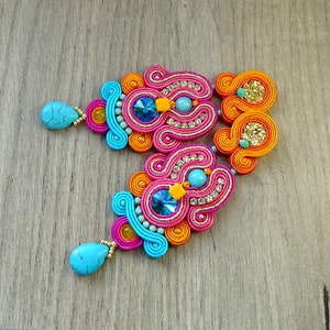 May include: A pair of colorful earrings with turquoise and pink accents. The earrings are made of woven thread and feature a large, sparkly blue stone in the center.