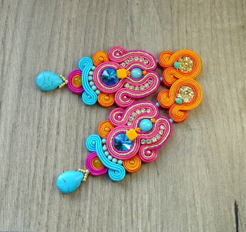 May include: A pair of colorful earrings with turquoise and pink accents. The earrings are made of woven thread and feature a large, sparkly blue stone in the center.