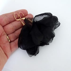 Könnte beinhalten: Ein Paar schwarze Stoffblumen-Ohrringe mit goldfarbener Hardware. Die Ohrringe haben ein zartes, geschichtetes Blütenblatt-Design. Goldfarbene Ohrhaken und gewundene Akzente verleihen den floralen Ohrringen Eleganz.