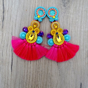 Pode incluir: Um par de brincos coloridos com borlas rosa, detalhes de contas azuis e amarelas e grandes strass amarelos.