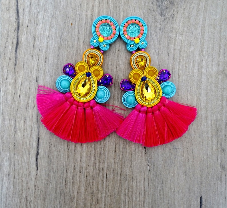 Pode incluir: Um par de brincos coloridos com borlas rosa, detalhes de contas azuis e amarelas e grandes strass amarelos.