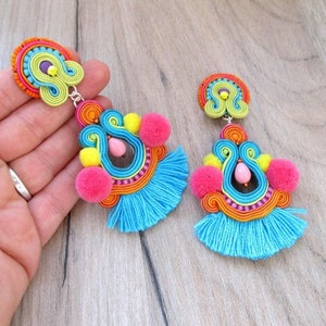 A pair of colorful dangle earrings with blue tassels, pink pom poms, and yellow pom poms. The earrings are made with a variety of colorful threads and beads.