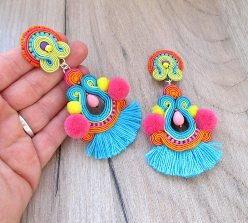 A pair of colorful dangle earrings with blue tassels, pink pom poms, and yellow pom poms. The earrings are made with a variety of colorful threads and beads.