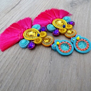 Pode incluir: Um par de brincos coloridos com borlas rosa, franjas de contas azuis e amarelas e detalhes turquesa e roxos.