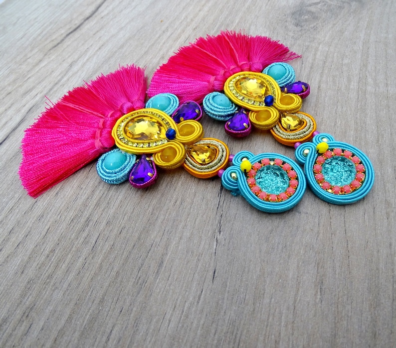 Pode incluir: Um par de brincos coloridos com borlas rosa, franjas de contas azuis e amarelas e detalhes turquesa e roxos.