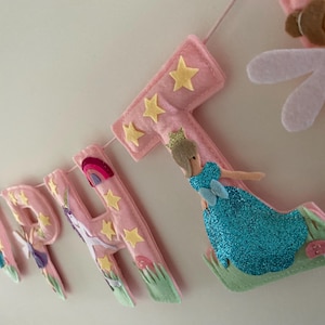 K&ouml;nnte beinhalten: Ein Filzbanner, das HAPPY BIRTHDAY in rosa Buchstaben buchstabiert. Jeder Buchstabe ist mit gelben Sternen, gr&uuml;nem Gras und kleinen Pilzen verziert. Eine Figur in einem blau glitzernden Kleid mit Krone ist am Buchstaben T befestigt, samt einer Fee.