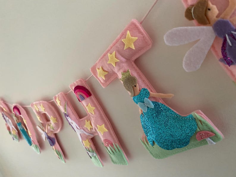 K&ouml;nnte beinhalten: Ein Filzbanner, das HAPPY BIRTHDAY in rosa Buchstaben buchstabiert. Jeder Buchstabe ist mit gelben Sternen, gr&uuml;nem Gras und kleinen Pilzen verziert. Eine Figur in einem blau glitzernden Kleid mit Krone ist am Buchstaben T befestigt, samt einer Fee.