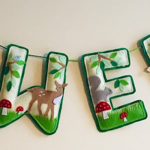 Könnte beinhalten: Ein Filzbanner mit dem Namen "OWEN" in grünen Buchstaben. Jeder Buchstabe hat ein anderes Waldtier- und Blattdesign.