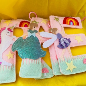 K&ouml;nnte beinhalten: Drei hellrosa Filzbuchstaben, die das Wort RAE bilden, verziert mit einem wei&szlig;en Einhorn, einer Figur in einem blaugr&uuml;nen Kleid, einer Fee mit violetten Fl&uuml;geln, Regenb&ouml;gen, gelben Sternen, gr&uuml;nem Gras und kleinen rosa Pilzen.