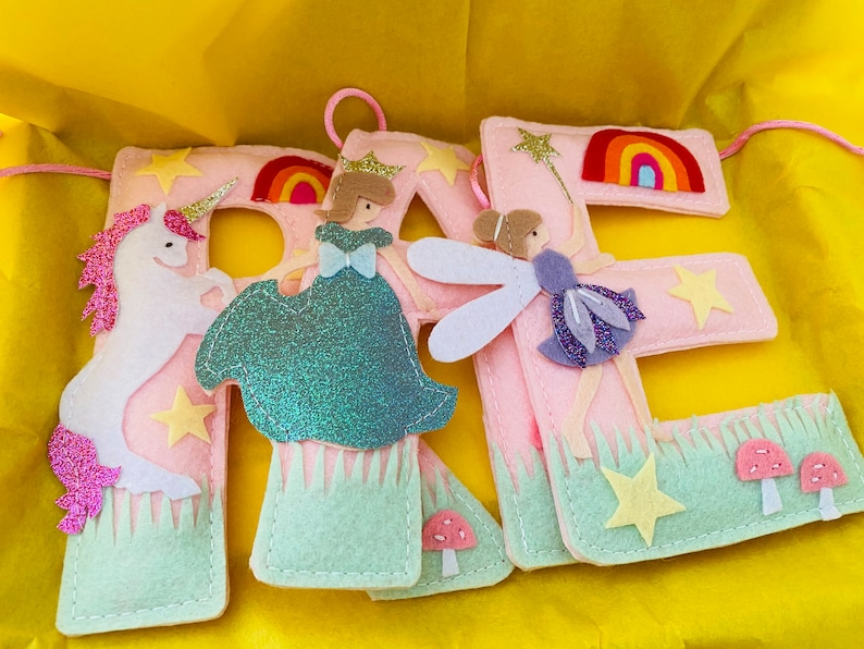K&ouml;nnte beinhalten: Drei hellrosa Filzbuchstaben, die das Wort RAE bilden, verziert mit einem wei&szlig;en Einhorn, einer Figur in einem blaugr&uuml;nen Kleid, einer Fee mit violetten Fl&uuml;geln, Regenb&ouml;gen, gelben Sternen, gr&uuml;nem Gras und kleinen rosa Pilzen.