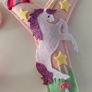 K&ouml;nnte beinhalten: Ein weicher Filzbuchstabe Y in Hellrosa, verziert mit einem wei&szlig;en Einhorn mit goldenem Glitzerhorn und violetter Glitzerm&auml;hne. Der Buchstabe enth&auml;lt gelbe Filzsterne, einen kleinen Regenbogen und eine gr&uuml;ne Basis mit einem kleinen rosa Pilz.