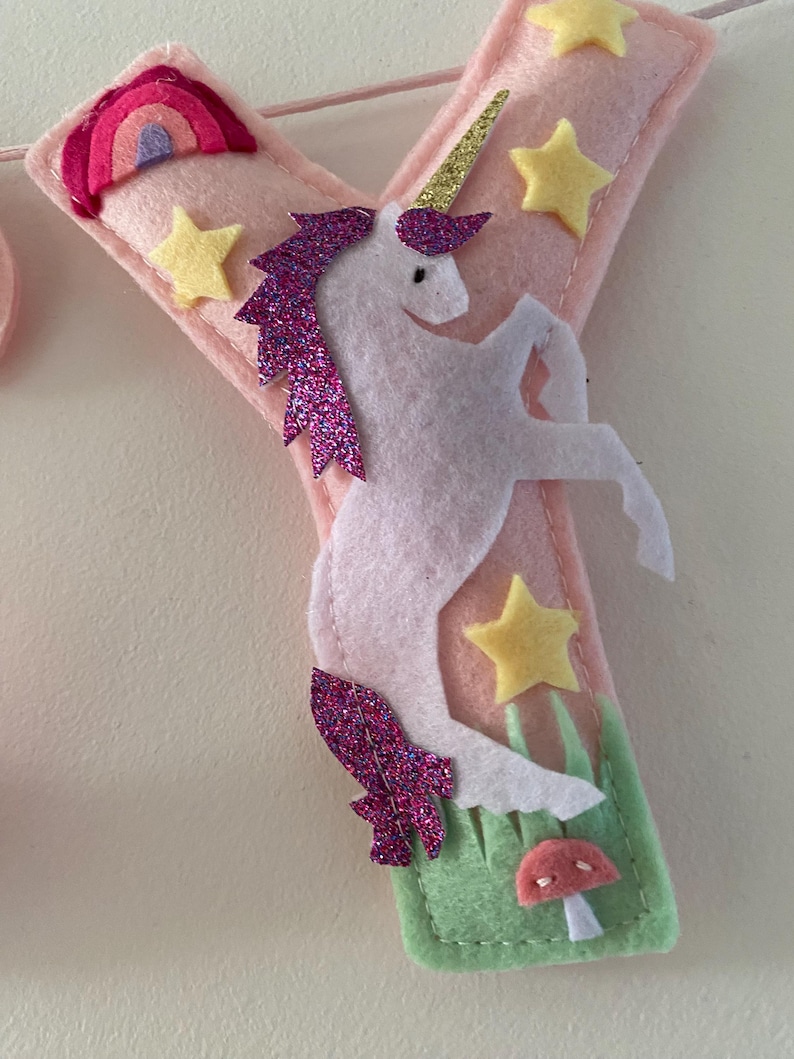 K&ouml;nnte beinhalten: Ein weicher Filzbuchstabe Y in Hellrosa, verziert mit einem wei&szlig;en Einhorn mit goldenem Glitzerhorn und violetter Glitzerm&auml;hne. Der Buchstabe enth&auml;lt gelbe Filzsterne, einen kleinen Regenbogen und eine gr&uuml;ne Basis mit einem kleinen rosa Pilz.